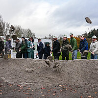 Auf dem Bild sind viele, darunter auch Oberbürgermeister Peter Boch zu sehen, die mit Schaufeln Sand in die Luft werden als symbolischer Spatenstich für den Skatepark im Enzauenpark.