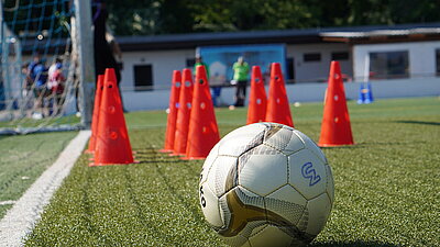 Ein weißer Fußball liegt vor 9 roten Kegeln auf dem Rasen.
