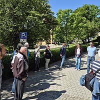 Eine Gruppe von Menschen verschiedenen Alters, die auf einem Parkplatz in einem Kreis stehen. Im Umfeld sind Bäume zu sehen und die Sonne scheint. 