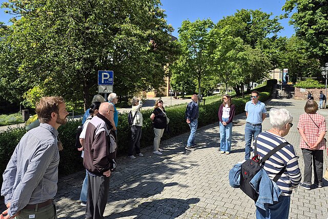 Eine Gruppe von Menschen verschiedenen Alters, die auf einem Parkplatz in einem Kreis stehen. Im Umfeld sind Bäume zu sehen und die Sonne scheint. 