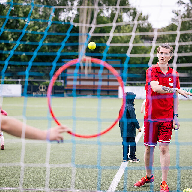 Das Foto ist hinter einem Fußballtor aufgenommen, sodass man das Netz des Fußballtors über das gesamte Foto sieht. Davor wirft ein Mann einen Ball in Richtung eines Reifens. - copyright:Stadt Pforzheim