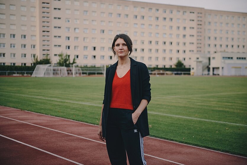 Frau mit dunklen kurzen Haaren steht vor einem Wohnblock auf einen Sportplatz. - copyright: Jacintha Nolte - Ersteller: Jacintha Nolte