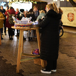 Eine Frau steht an einem Stehtisch, auf dem verschiedene Körbe stehen. Im Hintergrund sind beleuchtete Hütten und Besucher:innen des Weihnachtsmarkt zu sehen. - copyright:miteinanderleben e.V.