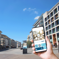 Die Pforzheimer Innenstadt. Im Vordergrund hebt eine Hand ein Handy, im Hintergrund ist eine Informationsstele zu sehen