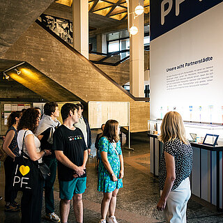 Ungefähr zehn Personen schauen sich eine beleuchtete Vitrine unter einer Überschrift: Unsere acht Partnerstädte an. - copyright:Stadt Pforzheim