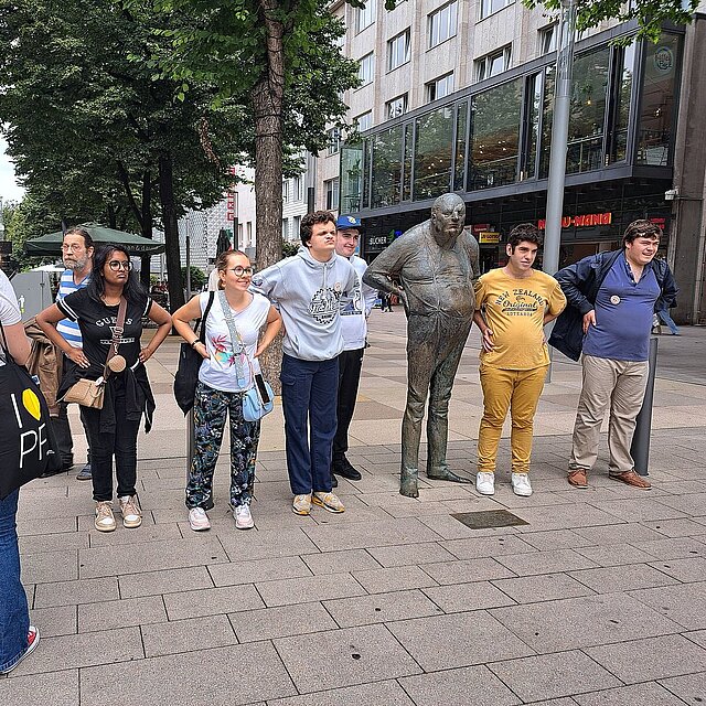 Neben der Statue "der Dicke" stehen rechts zwei junge Männer und links zwei junge Männer und zwei junge Frauen, die die gleiche Körperhaltung der Statue eingenommen haben. "Der Dicke" hat beide Hände hinten aufs Becken gelegt und streckt den Bauch nach vorne und schaut nach schräg vorne. - copyright:Stadt Pforzheim