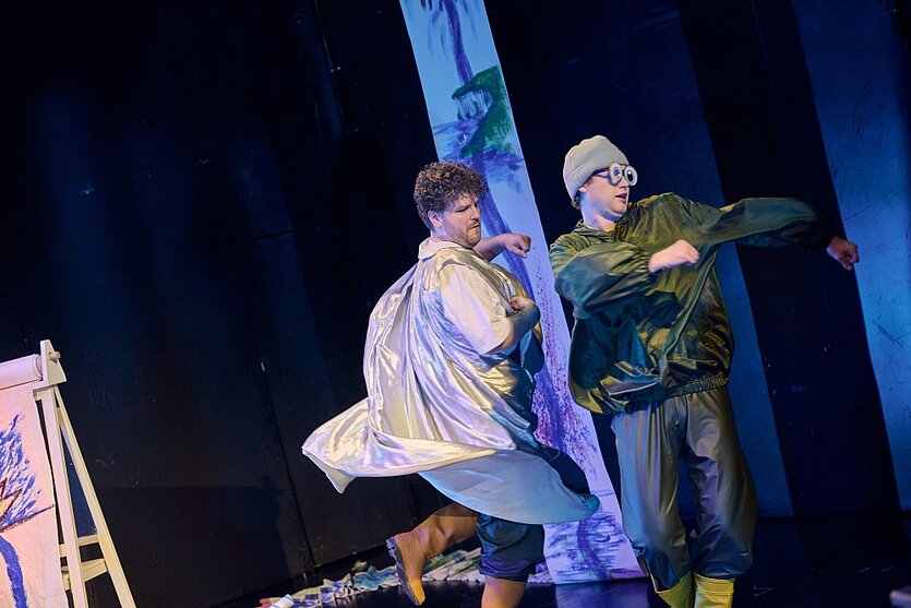 Zwei Personen stehen auf einer Bühne und bewegen sich dynamisch, als würden sie tanzen oder spielen, umgeben von farbigem Bühnenlicht. Im Hintergrund sind bemalte Leinwände und Requisiten zu sehen. - copyright: Stadttheater Pforzheim - Ersteller: Stadttheater Pforzheim