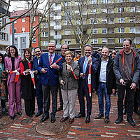 Viele Personen stehen nebeneinander auf einem Platz und halten je ein Stück rot-weißes Absperrband in den Händen