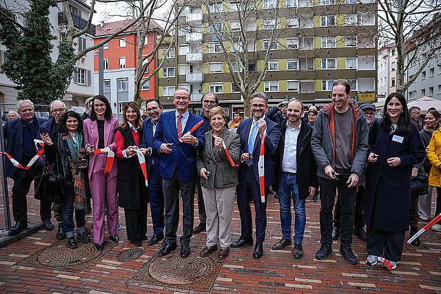 Viele Personen stehen nebeneinander auf einem Platz und halten je ein Stück rot-weißes Absperrband in den Händen