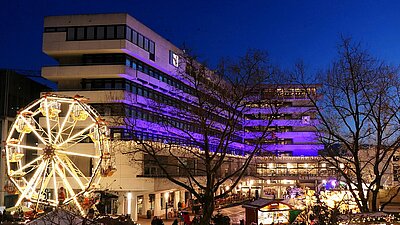 Auf dem Foto ist das Pforzheimer Rathaus zu sehen, das bei Dämmerung lila beleuchtet ist. Vor dem Rathaus auf dem Marktplatz ist der beleuchtete Weihnachtsmarkt zu sehen. Das Foto wurde von einer höheren Position aufgenommen.