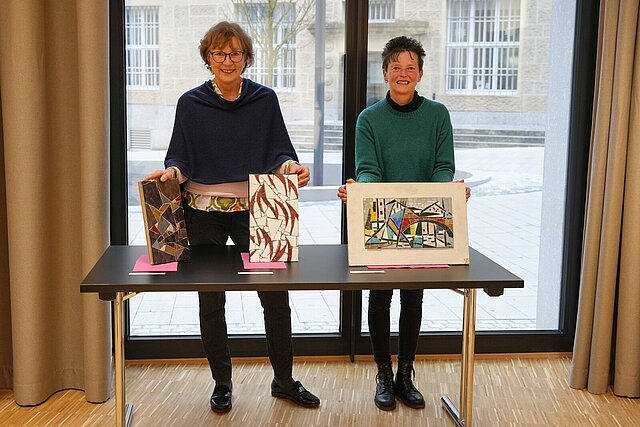 Zwei Frauen stehen hinter einem Tisch und halten drei Mosaik-Werke in den Händen