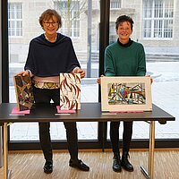 Zwei Frauen stehen hinter einem Tisch und halten drei Mosaik-Werke in den Händen