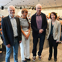 Ein Gruppenbild mit zwei Männern und zwei Frauen vor einer Treppe. Der Blick führt in den Saal mit Menschen, die sich an verschiedenen Ständen über Berufe informieren. 