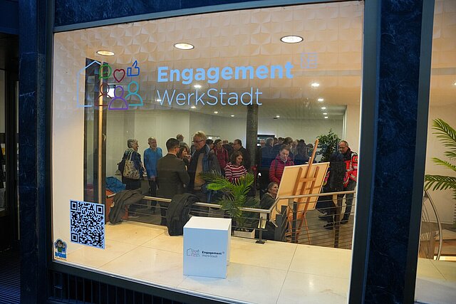 Ein Schaufenster von Außen, im Inneren sind mehrere Personen zu sehen.