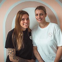 farbiges Bild von zwei Mitarbeiterinnen der Großtagespflege vor einer Regenbogenwand