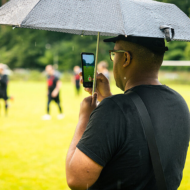 Ein Zuschauer macht Filmaufnahmen von einem Fußballspiel unter einem schwarzen Regenschirm. - copyright:Stadt Pforzheim