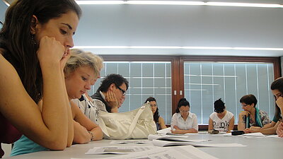 Farbfoto: Menschen verschiedenen Alters sitzen gemeinsam an einem Tisch und lesen.