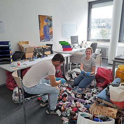 Ein Mann und eine Frau sitzen vor einem großen Haufen Socken. Sie tragen blaue Handschuhe und halten eine Socke in der Hand. - copyright:Stadt Pforzheim