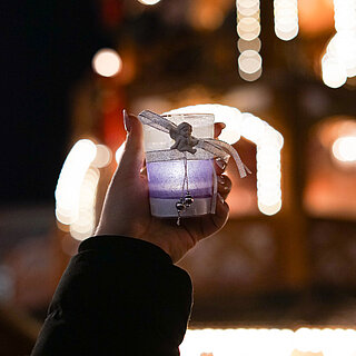 Auf dem Foto ist ein lila Windlicht zu sehen. Auf dem Windlicht ist eine Engelsfigur befestigt und mit einem Band umwickelt, an dem zwei Perlen hängen. Das Windlicht wird von einem Arm hochgehalten. Im verschwommenen Hintergrund ist ein stark beleuchtetes Gebäude zu sehen. - copyright:miteinanderleben e.V.