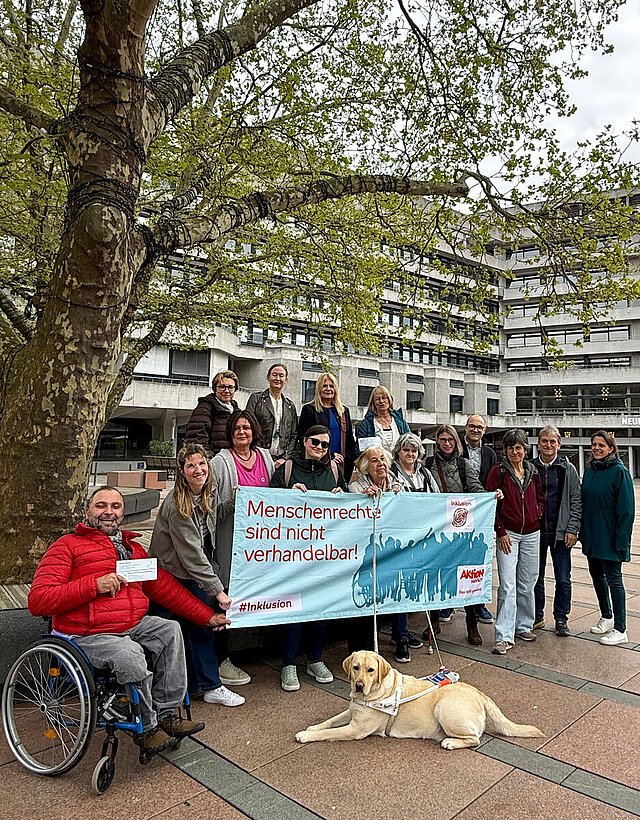 Gemeinsam mit dem Planungsteam stehen die Inklusionsbeauftragten der Stadt Pforzheim und des Enzkreises vor einem Baum auf dem Marktplatz der Stadt.