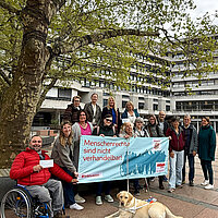 Gemeinsam mit dem Planungsteam stehen die Inklusionsbeauftragten der Stadt Pforzheim und des Enzkreises vor einem Baum auf dem Marktplatz der Stadt.