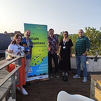 5 Personen auf einer Dachterrasse neben einem grünen Aufsteller.