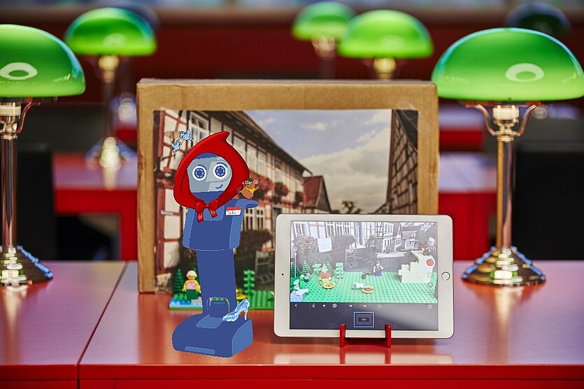 Auf einem Tisch stehen ein Tablet und eine kleine Szene aus Spielzeugfiguren, daneben eine stilisierte, blau-rote Figur im Vordergrund. Im Hintergrund sind grüne Schreibtischlampen und ein gerahmtes Bild einer Fachwerkstraße zu sehen. - copyright: Stadtbibliothek Pforzheim - Ersteller: Stadtbibliothek Pforzheim