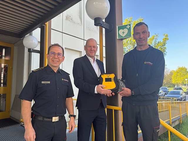 Feuerwehrkommandant Sebastian Fischer, Erster Bürgermeister Dirk Büscher und Notfallsanitäter sowie Praxisanleiter Stephan Bürkle (von links) stellen den neuen, öffentlich zugänglichen AED an der Hauptfeuerwache vor. Sie stehen dabei vor dem Eingang zur Hauptfeuerwache.
