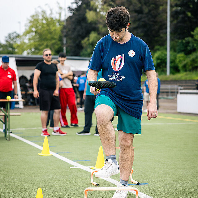 Ein junger Mann hält in der linken Hand einen Schläger auf dem er einen Ball balanciert. Gleichzeitig steigt er über ein Hindernis. - copyright:Stadt Pforzheim