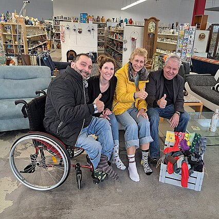 In einem Laden sind ein Rollstuhlfahrer, zwei Frauen und ein Mann zu sehen. Sie sitzen hinter einen Box mit bunten Socken. Sie tragen keine Schuhe, aber verschiedene Socken. - copyright:Stadt Pforzheim