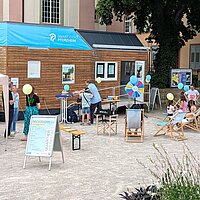 Ein Tiny House steht auf einem Platz, davor sind Kinder und Erwachsene zu sehen.