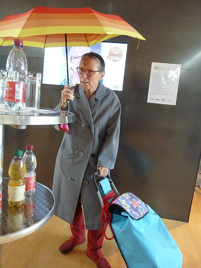 Farbfoto: ältere Frau steht mit gespanntem Regenschirm und Einkaufstasche und guckt auf Wasserflaschen auf einem Stehtisch