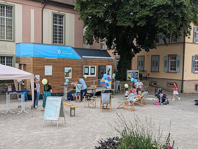 Eine kleines Haus steht auf einem Platz. Erwachsene und Kinder stehen davor