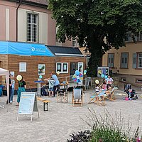Eine kleines Haus steht auf einem Platz. Erwachsene und Kinder stehen davor