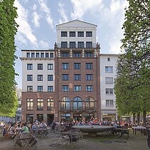 Farbfoto: Sedanplatz mit "Haus am Sedanplatz", sitzenden Menschen und dem Brunnen in der Mitte. - copyright:Günter Beck