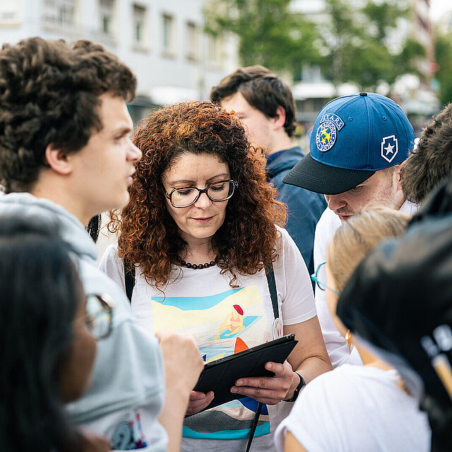 Eine Frau mit rotbraunen Locken und Brille hält ein Tablet in der Hand und schaut darauf. Mehrere Personen stehen im Kreis um die Frau und schauen auf das Tablet - copyright:Stadt Pforzheim