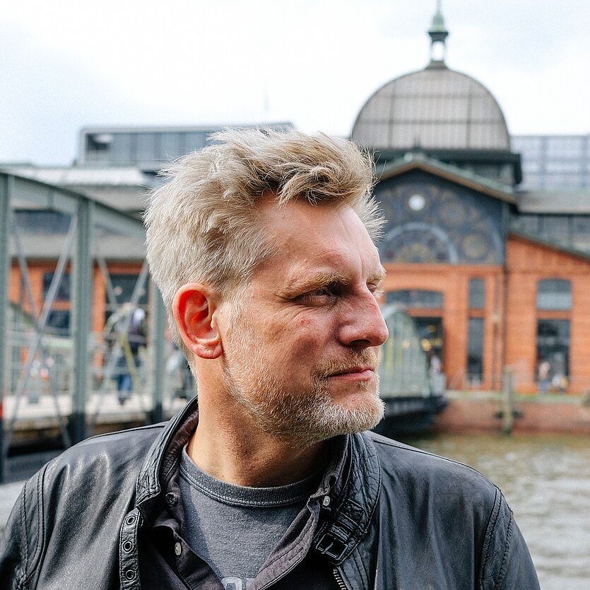 Porträt eines Mannes mit kurzem blondem Haar und Bart, der seitlich blickt und eine schwarze Lederjacke trägt. Im Hintergrund ein Fluss mit Brücke sowie ein Gebäude mit Kuppeldach. - copyright: Elena Gohdes - Ersteller: Elena Gohdes