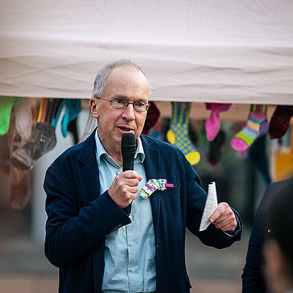 Ein Mann mit hellblauen Hemd und dunkelblauen Sakko hält ein Mikrophon in der Hand und spricht. In der anderen Hand hält er einen Zettel. Am Sakko ist eine kleine, bunte, gestrickte Socke mit rosa Wäscheklammer befestigt. - copyright:Paul Hoffer