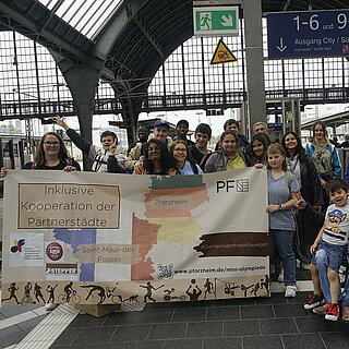 Eine Gruppe von etwa 20 Personen steht auf einem Gleis am Bahnhof. Im Vordergrund ist ein Banner mit der Aufschrift "Inklusive Kooperation der Partnerstädte" zu sehen. - copyright:Stadt Pforzheim