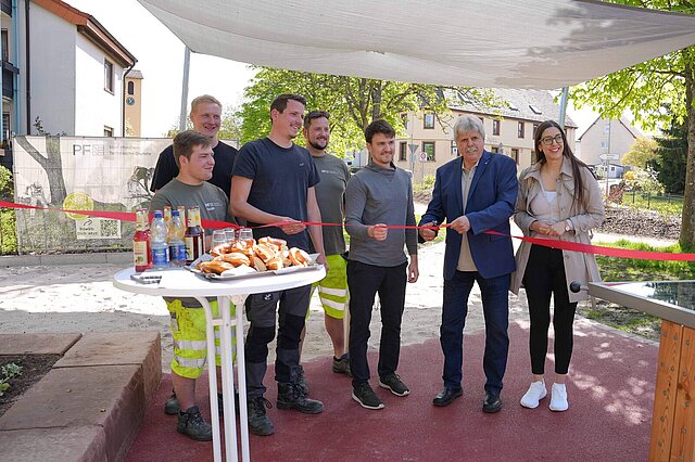 Viele Personen stehen hinter einem roten Band, zwei von ihnen scheiden das Band durch. Im Hintergrund ist ein Spielplatz zu sehen.
