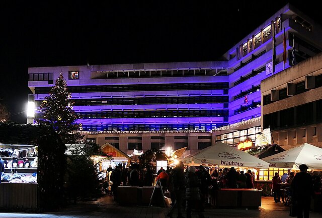 Auf dem Foto ist das Pforzheimer Rathaus zu sehen, das bei Dämmerung lila beleuchtet ist. Vor dem Rathaus auf dem Marktplatz ist der beleuchtete Weihnachtsmarkt zu sehen. Das Foto wurde auf dem Weihnachtsmarkt aufgenommen. - copyright:Stadt Pforzheim