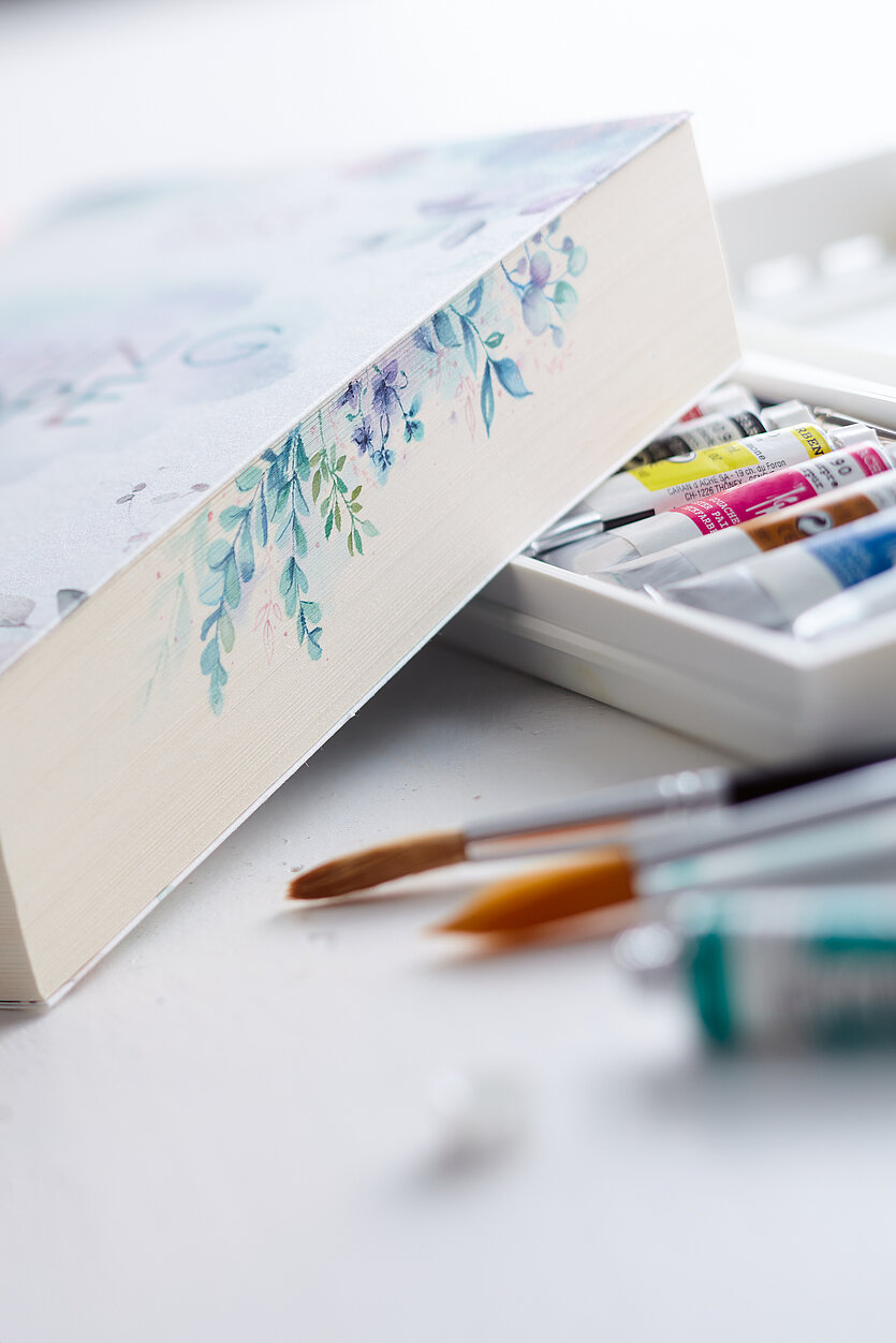 Nahaufnahme einer dekorativ gestalteten Box mit floralem Muster, die leicht geöffnet ist und Aquarellfarben enthält. Im Vordergrund liegen unscharf mehrere Pinsel auf einem hellen Tisch. - copyright: TOPP-Verlag - Ersteller: TOPP-Verlag