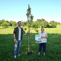 Ein Mann und eine Frau stehen neben einem kleinen Apfelbaum auf einer großen Wiese. Im Hintergrund sind Bäume zu sehen.