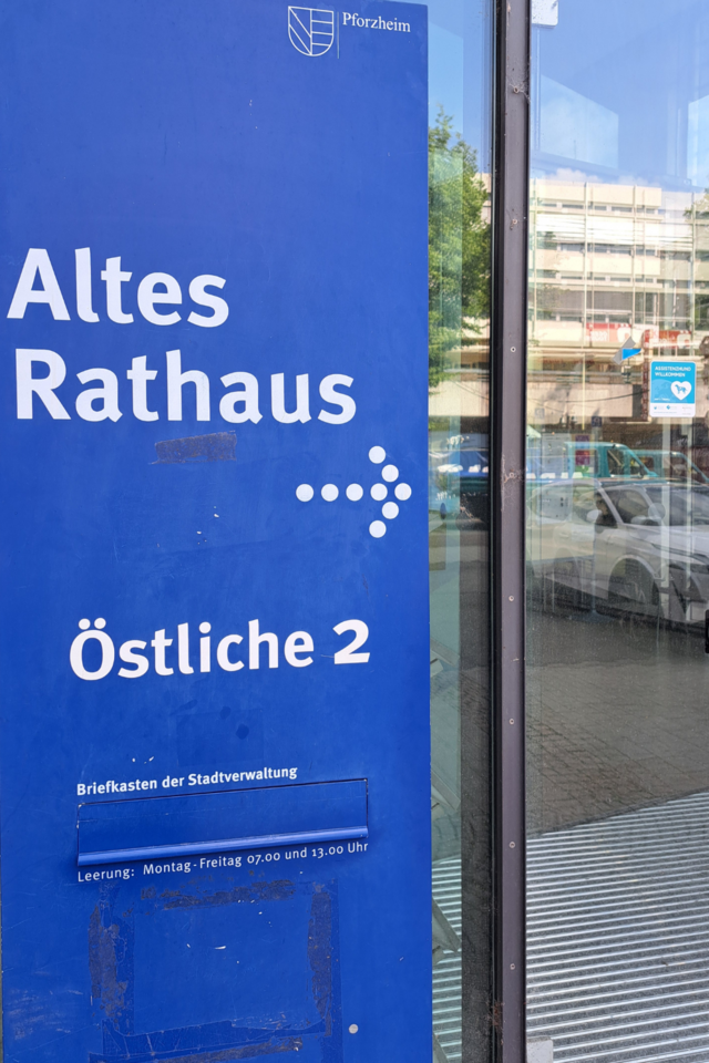 Zu sehen ist links eine blaue Stehle mit der weißen Aufschrift "Altes Rathaus, Östliche 2" und einem Pfeil der nach rechts zeigt. Rechts neben der blauen Stehle ist eine Glastür mit dem Aufkleber "Assistenzhunde Willkommen" - copyright:Stadt Pforzheim